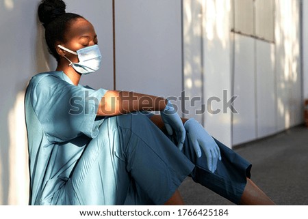 Tired exhausted female african scrub nurse wears face mask blue uniform gloves sits on hospital floor. Depressed sad black ethic doctor feels fatigue burnout stress, lack of sleep, napping at work.