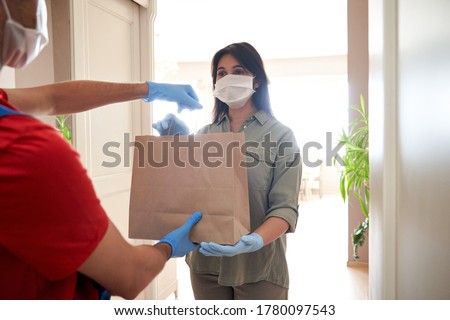 Indian woman customer wearing face mask and gloves taking delivery paper eco bag from man courier holding grocery food package delivering supermarket takeaway order standing at home. Safe delivery.