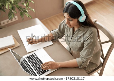 Indian girl student wear headphones learning online watching webinar class looking at laptop computer elearning remote lesson making notes or video calling virtual conference meeting teacher at home.
