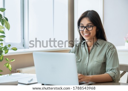 Happy indian young adult woman wearing glasses using pc laptop computer working studying at home office sitting at table. Happy female professional freelancer learning watching online webinar training