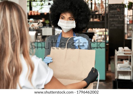 African American female cafe worker wears face mask and gloves giving takeaway food bag to customer. Mixed race waitress holding takeout order standing in coffee shop restaurant with take away client.