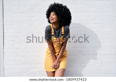 Happy stylish African American young woman wears yellow trendy sundress with Afro hair laughing looking away standing against white brick wall outdoor background. Smiling black hipster woman portrait