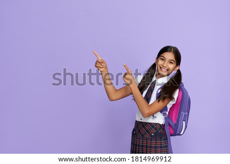 Happy indian kid primary elementary school girl with backpack wearing school uniform pointing fingers aside at copy space advertising products or services for pupils isolated on violet background.