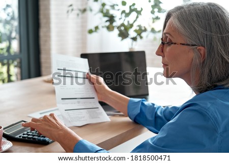 Senior mature business woman holding paper bill using calculator, old lady managing account finances, calculating money budget tax, planning banking loan debt pension payment sit at home office table.