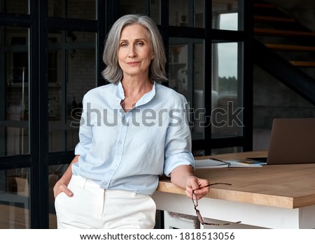 Photo of Confident stylish european mature middle aged woman standing at workplace. Stylish older senior businesswoman, 60s gray-haired lady executive leader manager looking at camera in office, portrait.