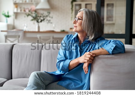 Calm relaxed mature older woman relaxing sitting on couch at home. Peaceful middle aged grey-haired lady resting on sofa in modern living room enjoying lounge and no stress, looking away, thinking.