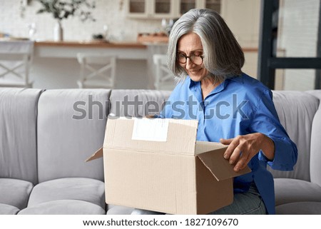 Smiling older adult mature woman customer unpacking parcel concept sitting at home on couch. Happy senior middle aged lady opening online store order receiving gift in postal delivery shipping box.