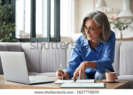 Happy stylish mature old woman remote working from home distance office on laptop taking notes. Smiling 60s middle aged business lady using computer watching webinar sit on couch writing in notebook.