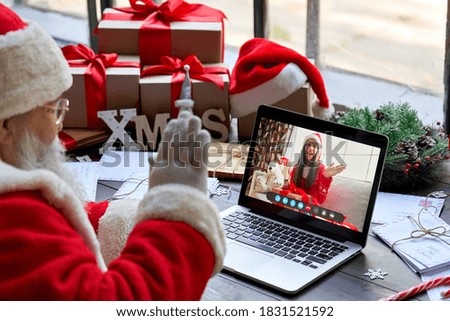 Over shoulder view of Santa Claus video calling kid girl on laptop greeting child by webcam talk open Christmas gift box in virtual online chat meeting using computer sit at workshop table on xmas.