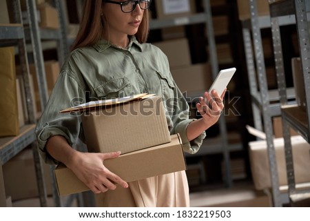 Female warehouse worker manager, small stock business owner holding phone and retail package parcel boxes checking commercial shipping delivery order on smartphone using mobile app technology.