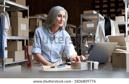 Mature female fashion seller using computer checking ecommerce clothing store orders. Older middle aged business woman entrepreneur working on laptop preparing online shipping delivery parcels boxes.