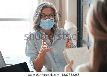 Old middle aged female mentor, teacher, hr manager wearing face mask training young worker intern, teaching student, interviewing job seeker at meeting in office. Social distance and safety at work.