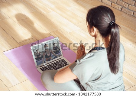 Young woman meditating watching live online tv pilates group class tutorial on laptop computer at home doing yoga virtual training fitness workout meditation exercise stream. Over shoulder screen view