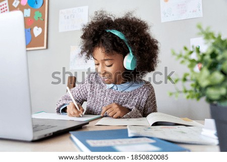 Cute smiling african school kid girl wearing headphones virtual distance learning online listening remote education digital class doing homework studying at home classroom sitting at desk with laptop.