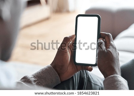 African american man holding smart phone with mockup white blank display, empty screen for app ads sitting on couch at home. Mobile applications technology concept, over shoulder close up view.