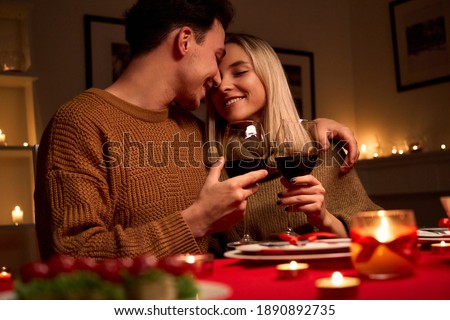 Happy young couple in love hugging holding glasses, drinking wine, celebrating Valentines day dining at home together, having romantic dinner date with candles sitting at table, embracing and bonding.