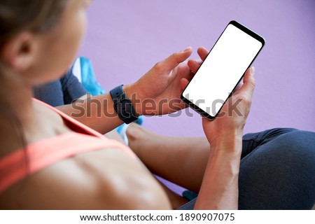 Over shoulder closeup view of fit sporty woman yoga teacher sit on mat holding phone mock up screen video calling, watching online workout training class video, using fitness app at home in gym.