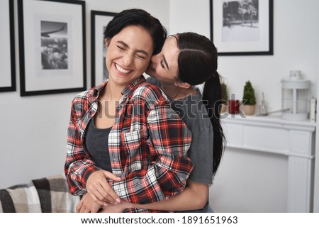 Happy lesbian lgbtq couple in love cuddling, laughing, whispering on ear having fun standing at home. Two stylish cool diverse pretty affectionate women hugging, bonding. Lgbt relationship concept