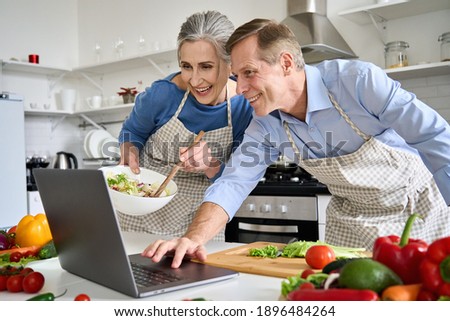 Happy old middle aged 50s couple using laptop computer preparing healthy food diet vegetable salad at home together, searching recipes, ordering shopping online, watching cooking class in kitchen.