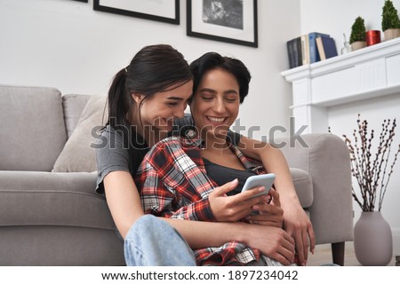 Young happy lesbian lgbtq couple or friends holding smartphone using mobile phone at home, doing online shopping in ecommerce app together, having fun checking social media applications together.