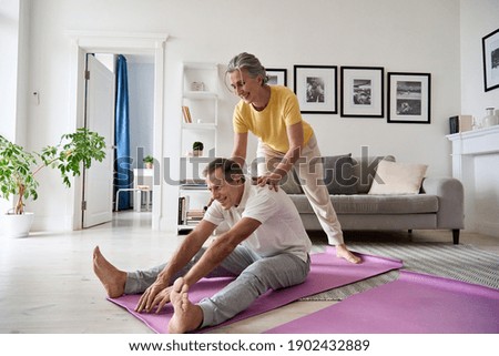 Fit active retired middle aged wife helping senior husband doing stretching exercise at home. Happy healthy older senior 60s couple enjoying fitness sport training workout together in apartment.