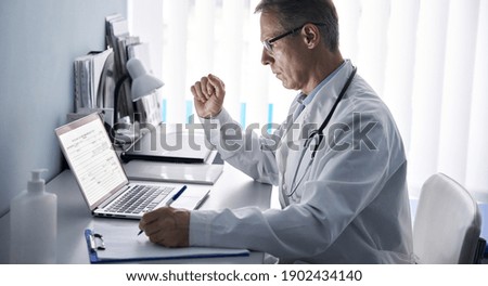 Serious mature old doctor physician using laptop tech in hospital office. Senior middle aged male gp checking patient clinical registration form, elearning working looking at computer sitting at desk.