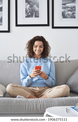 Smiling latin teenage girl holding smartphone using mobile apps technology at home. Happy hispanic young woman texting, checking online social media applications sitting on couch in apartment.