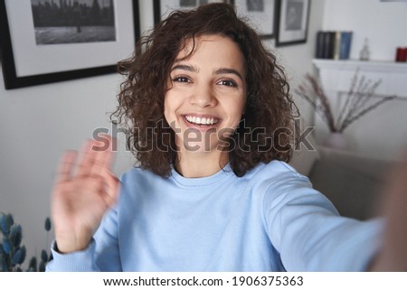 Happy pretty hispanic gen z teen girl vlogger holding smartphone waving hand, talking to camera shooting vlog, making video call at home talking by virtual mobile video call chat app. Webcam view.