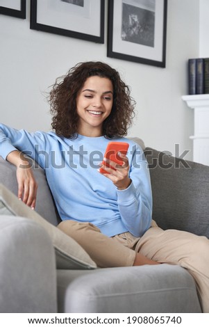 Happy young woman watching video looking at smartphone relaxing on couch, smiling hispanic teen girl enjoying using online mobile apps for education or entertainment on cell phone at home.