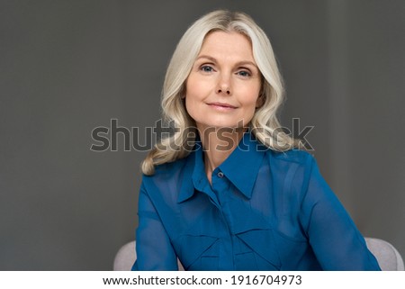 Sophisticated elegant beautiful 50s attractive middle aged smiling woman model sitting in chair looking at camera at home. Portrait of gorgeous confident mature older blond hair lady indoors.