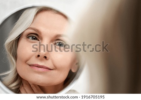 Photo of Happy 50s middle aged woman model touching face skin looking in mirror reflection. Smiling mature old lady pampering, healthy moisturized skin care, aging beauty, skincare treatment cosmetics concept.