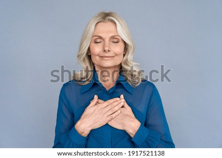Happy mindful thankful middle aged old woman holding hands on chest meditating with eyes closed isolated on grey background feeling no stress, gratitude, mental health balance, peace of mind concept.