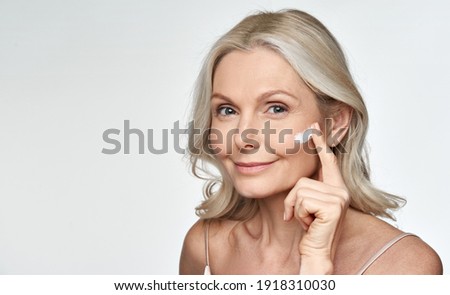 Smiling 50s middle aged mature older woman applying facial cream on face looking at camera isolated on white background. Anti age healthy dry skin care beauty therapy concept, old skincare treatment