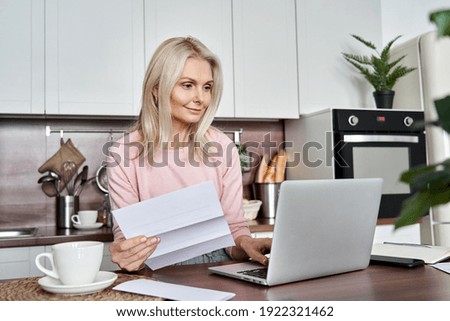 Middle aged mature woman holding paper bill or letter using laptop computer at home for making online payments on website, calculating financial taxes fee cost, reviewing bank account, loan rates.