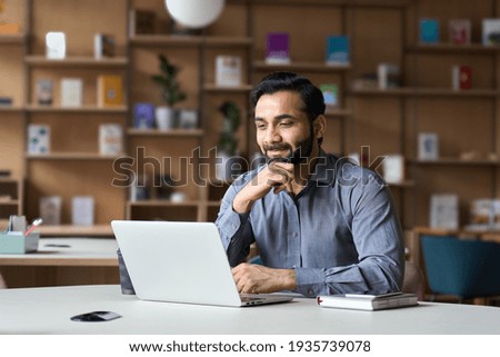 Smiling indian businessman working on laptop in modern office lobby space. Young indian student using computer remote studying, watching online webinar, zoom virtual training on video call meeting.