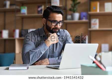 Young serious indian professional business man, focused ethnic male student wearing glasses working on laptop, remote studying using computer looking at screen watching seminar webinar at home office. Foto stock © 