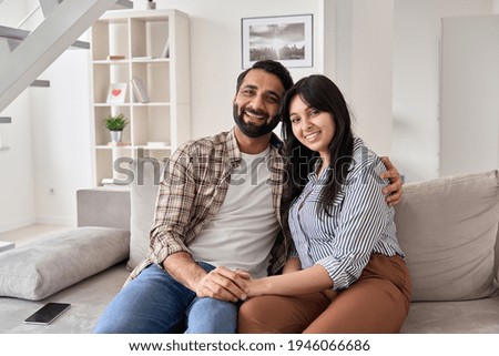 Happy young indian couple real estate buyers hugging sitting on couch at home looking at camera. Smiling husband and wife new homeowners embracing, enjoying own apartment purchase, portrait.