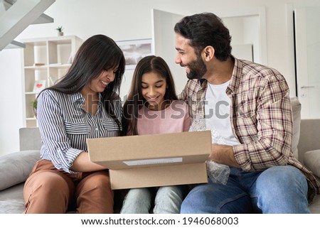 Happy indian family with child daughter unpacking parcel at home. Smiling parents and teen kid daughter opening postal box looking at gift in online shopping delivery package sitting on sofa together.