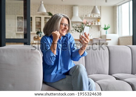 Great news. Happy surprised mature 60s aged woman super excited reacting to cell phone sms winning grandiose surprise. Feeling great happiness of receiving gift on sofa at home.