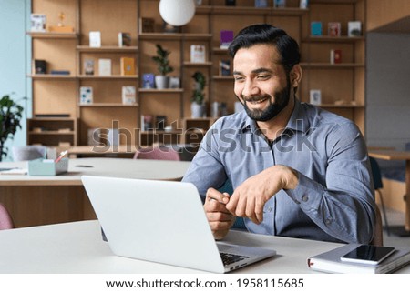 Happy smiling ethnical indian businessman having online virtual meeting call on laptop looking at screen sitting at table in coworking creative office, professional manager talking to employees.