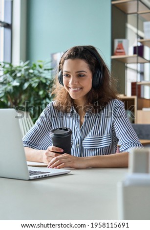 Young adult happy smiling Hispanic indian student wearing headphones talking online chat meeting holding coffee using laptop in virtual office. College female student learning remotely.