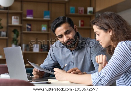 Two multiethnic professionals colleagues working together with laptop and papers in office. Indian male mentor and latin female young professional sitting in creative office space.