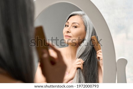 Senior attractive middle 50 years aged asian woman with gray hair looking at mirror reflection combing tangled gray hair. Alopecia hair loss prevention treatment after menopause advertising concept.