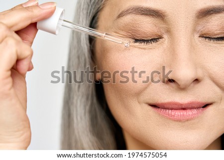 Closeup cut portrait of senior mature older Asian woman with closed eyes touching face eye contour with antiaging pipette serum essence oil. Anti wrinkle prevention eye skin care products concept.