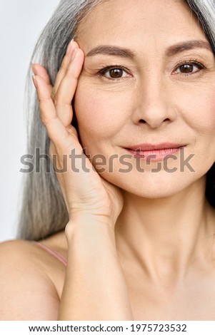 Vertical portrait of middle aged Asian woman's face with perfect skin. Older mature lady touching pampering face with hand. Advertising of cosmetology salon rejuvenating spa procedures skincare.
