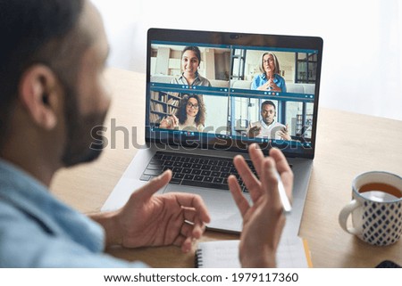 Latin indian businessman having virtual team meeting group call chatting with diverse people in customer support. Video conference call on computer with manager and employees. Over shoulder view.