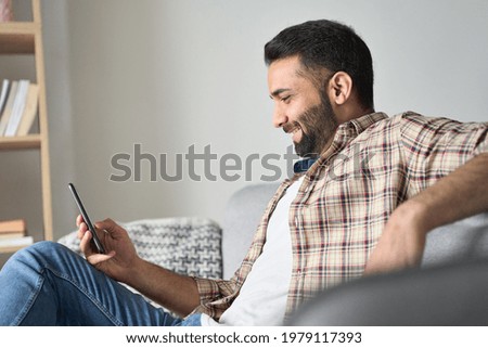 Young relaxed smiling indian man sitting on couch in modern living room looking at cellphone checking social media, surfing internet, ordering food delivery, using mobile apps at weekend at home.