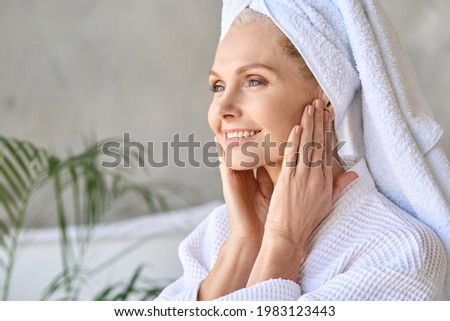 Happy smiling gorgeous middle aged woman wearing bathrobe and white towel touching face looking at window. Advertising of skin care spa wellness salon procedures concept. Closeup portrait.