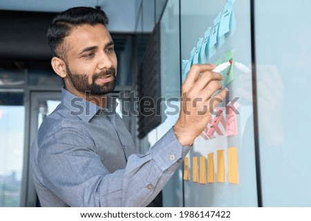 Young indian latin executive manager businessman ceo writing his strategy ideas on sticky notes on whiteboard managing professional organization business plan project presentation in corporate office.