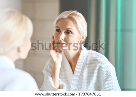 Gorgeous mid age adult 50 years old blonde woman standing in bathroom after shower touching face, looking at reflection in mirror doing morning beauty routine. Older dry skin care concept.